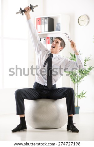 Successful businessman sitting in the office on pilates ball and doing exercise with dumbbells.
