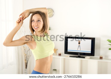 Close-up of a young woman in sports clothes, doing stretching exercises in the room, in front TV.