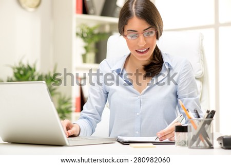 A young beautiful businesswoman sitting at an office desk in front of a laptop and writing in planner with the left hand.