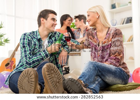 Young couple at a home party, sit on the floor, knocking the glasses toasting, in the background you can see another couple.