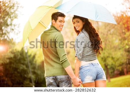 Rear view of a young couple in love with umbrellas while walking in the rain through the park holding hands and looking at camera other with a smile.