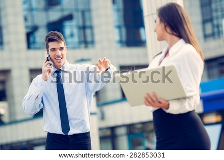 Young business partners standing in front of the building. Businessman talking on a mobile phone while his woman partner  working on a laptop.