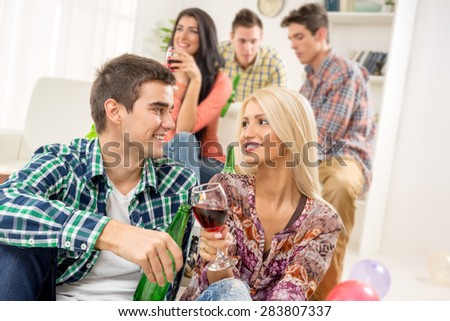 Young couple at a home party, sit on the floor, knocking the glasses toasting, in the background you can see friends.