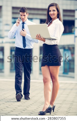 Young business partners standing in front of the building. Business woman working on a laptop while her partner use a mobile phone for business conversation.