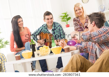 A small group of young people hang out at the house party, chatting with each other while their friend having fun playing acoustic guitar.