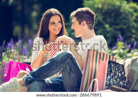 A young loving couple resting on a park bench after shopping, enjoying the sun while nibbling popcorn.