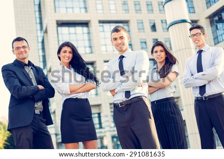 A small group of young business people standing in front of office building with arms crossed and with a smile on their faces looking at camera.