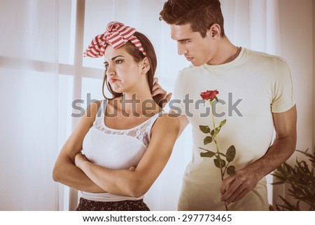Displeased beautiful girl standing not interested while he is cute boyfriend in love holding rose.