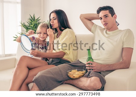 Young man sitting on sofa and watching football on tv. His wife and daughter having fun and make up.