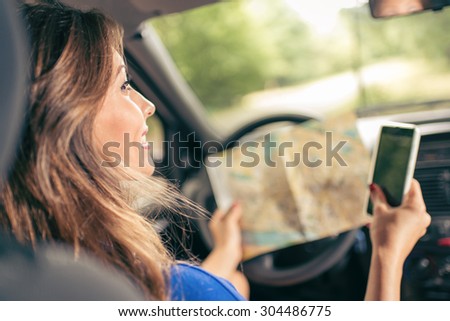 Young smiling beautiful woman sitting in car, holding map and smart phone. Rear view.