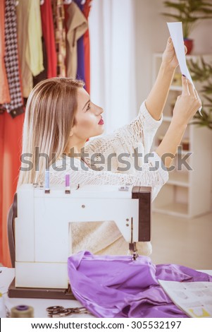 Young woman sits in front of the sewing machine and looking at sketches of clothing.