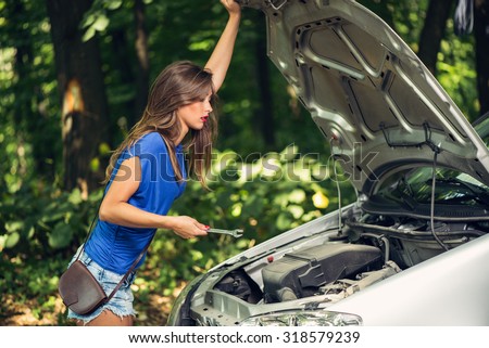 Beautiful young worried woman standing by car that broke down on the road in forest and holding wrench.