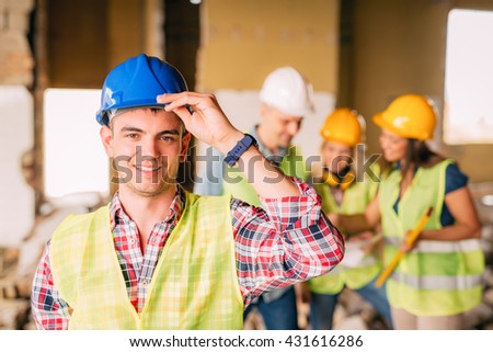Smiling young electrician holding helmet and looking at camera. His architect colleagues review plan in background at a construction site.