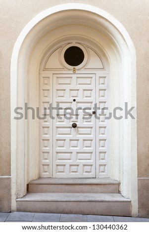 A modern single wooden door painted in white, with a doorknob. The entrance to the building has been placed sligthly receded and above from the street (hence the deep arched frame and the two steps).
