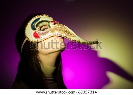 A girl wearing a venetian mask featuring a large nose, looking at camera. The shadow of her face (plus the mask\'s nose) is projected on the background. Illuminated using yellow and magenta spotlights.