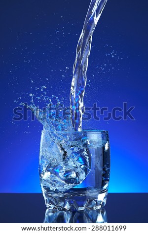 Pour fresh splash water on glass, blue background. Pitcher of water poured into a glass. Image refreshing water that is poured and produces splashes and drops of water. Cool healthy purity beverage.