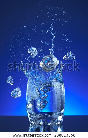 Pour fresh splash water on glass and ice cubes, blue background. Pitcher of water poured into a glass. Image refreshing water is poured and produces splashes and drops of water. Cool purity beverage.