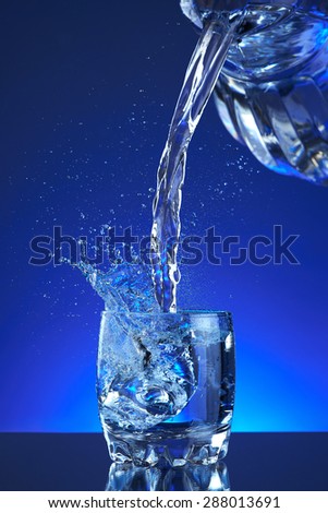 Pour fresh splash water on glass, blue background. Pitcher of water poured into a glass. Image refreshing water that is poured and produces splashes and drops of water. Cool healthy purity beverage.