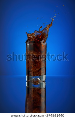 Refreshing splash of cola soft drink on a blue background. Liquid drink cola pouring into a glass with ice. Pour high speed beverage for promoting restaurant and bar. Closeup isolated design liquor.