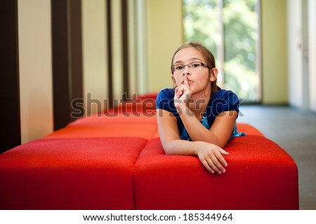 Little girl lifestyle shot in  a sunny room with her finger to her lips asking for quiet or silence laying on a red couch with an out of focus background and  room for copy