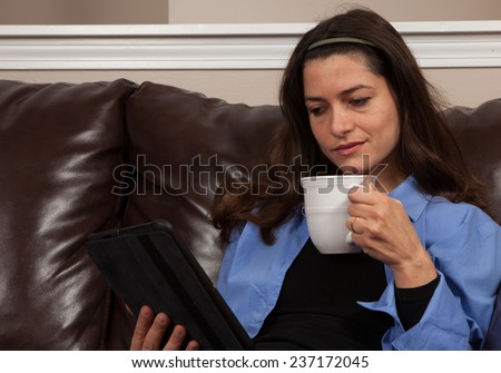 Happy young woman sitting alone on a couch with a tablet and mug on a brown leather couch with room for copy above