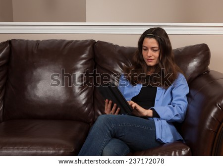 Happy young woman sitting alone on a couch with a tablet on a brown leather couch with room for copy above
