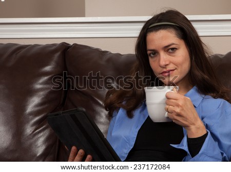 Happy young woman sitting alone on a couch with a tablet and mug on a brown leather couch with room for copy above