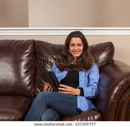 Happy young woman sitting alone on a couch with a tablet on a brown leather couch with room for copy above