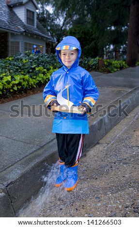 Young boy playing with toy boat in the rain wearing rain slickers and galoshes.