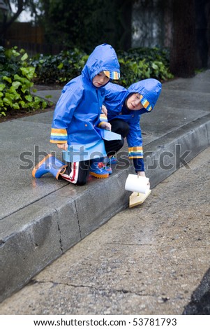 Young boys playing with toy boat in the rain wearing rain slickers and galoshes.