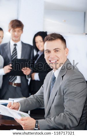 handsome young businessman happy smile. Sitting at the desk at office with group of business colleagues people in the background businesspeople working in team at meeting, man looking at camera