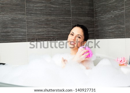 Beautiful smile woman washing shoulder with pink sponge in bath with foam, young girl bathing in bathtub, relaxing in bath  jacuzzi