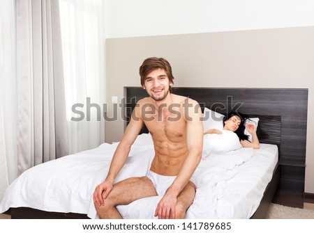 young couple in bedroom, happy smile man sitting, woman sleep lying in a bed with closed eyes