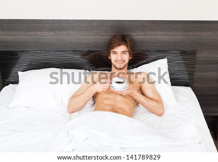 Handsome man lying on bed drink coffee, happy smile waking up in the morning hold cup tea, attractive guy smiling in bedroom at home, sexy young male model