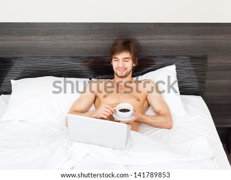 Handsome man lying on bed with laptop, happy smile drink coffee in the morning, attractive guy smiling in bedroom at home using computer, sexy young male model
