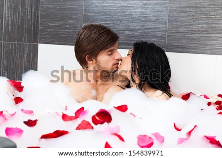 young couple kissing lying in jacuzzi, tub full of foam and red rose petals, concept of kiss romantic love