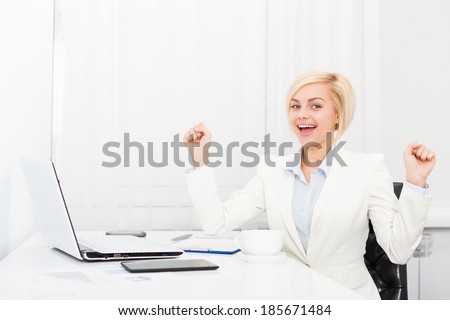 Business woman excited hold fist hands up raised arms sitting at modern office desk, surprised happy smile businesswoman success