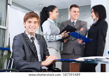 handsome young businessman happy smile. Sitting at the desk at office with group of business colleagues people in the background businesspeople working in team at meeting, man looking at camera