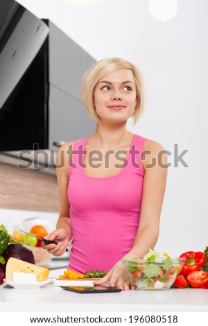 Blonde woman think looking up copy space, using a tablet computer to cook in modern kitchen, young girl fresh raw vegetables cooking at home smile dream