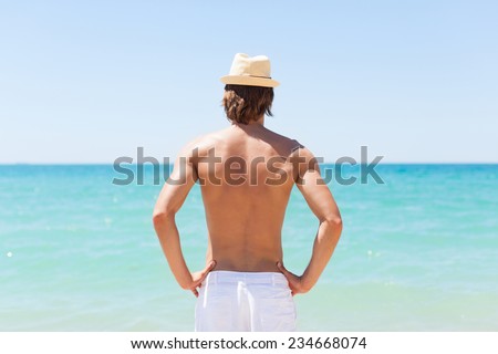 man on beach rear view guy wearing summer hat, standing back looking to sea blue sky horizon, vacation concept of freedom travel ocean