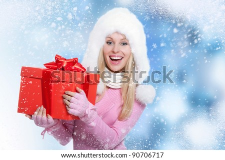 Christmas Girl. Winter new year woman hold gift box, happy smile woman wear warm fur hat scarf gloves holding, over abstract christmas ornament with wind blowing snow flake background
