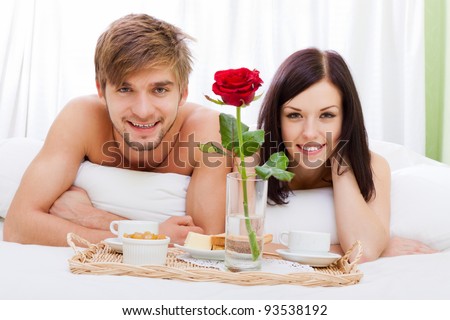 happy young couple eat breakfast in bed in morning with red rose flower, lovely couple lying in a bed, happy smile looking at camera, concept of honeymoon, valentine day