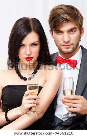 Portrait young couple with glass of champagne, wear evening dress red lips and suit with red bow, glamour vogue style, sensual looking at camera, sitting indoors