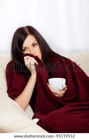 Ill woman covered with blanket holding cup of tea, sitting at home on sofa couch