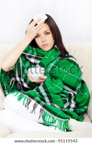 Ill woman covered with blanket, hold hand on forehead check temperature, holding cup of tea and pack of pills, sitting at home on sofa couch