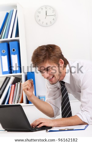 Successful excited young business man happy smile sitting at the desk hold raise arm hand up using computer, handsome businessman working on laptop typing, concept of success, victory, win