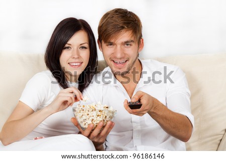 Couple watching tv in their living room at home, young happy smile man and woman eat popcorn, sitting on couch, sofa, hold remote control