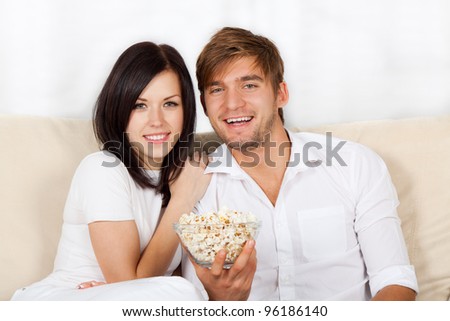 Couple watching tv in their living room at home, young happy smile man and woman eat popcorn, sitting on couch, sofa
