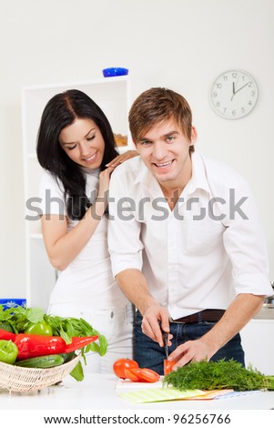 portrait of young lovely couple slicing tomato in their kitchen, man looking at camera woman embrace, hug happy smile