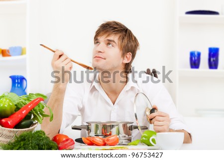 portrait of handsome young man hold spoon taste, pan with ready meal, soup, boiler in the kitchen, happy smile, think looking up